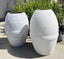 Two outdoor Sak' GFRC cylindrical outdoor planters with a white stucco texture. One is positioned upright while the other is on its side, showcasing the robust material and design.