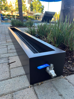A black fiberglass planter rain storage trough with a spigot, situated on a brick pathway with plants in the background.